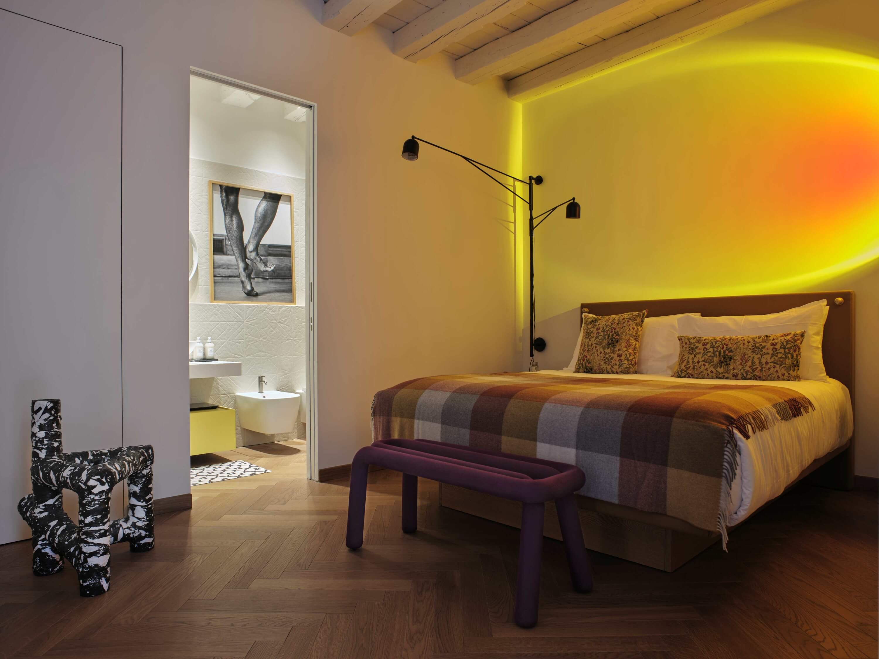 Camera da letto accogliente con bagno adiacente, letto con coperta a quadri, panchina e lampada da terra.